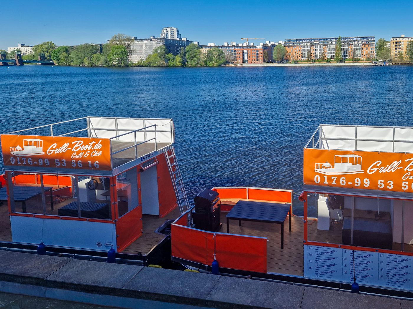 Zwei Grillboote von Grill-Boot.de mit Telefonnummer am Ufer vor der Berliner Skyline